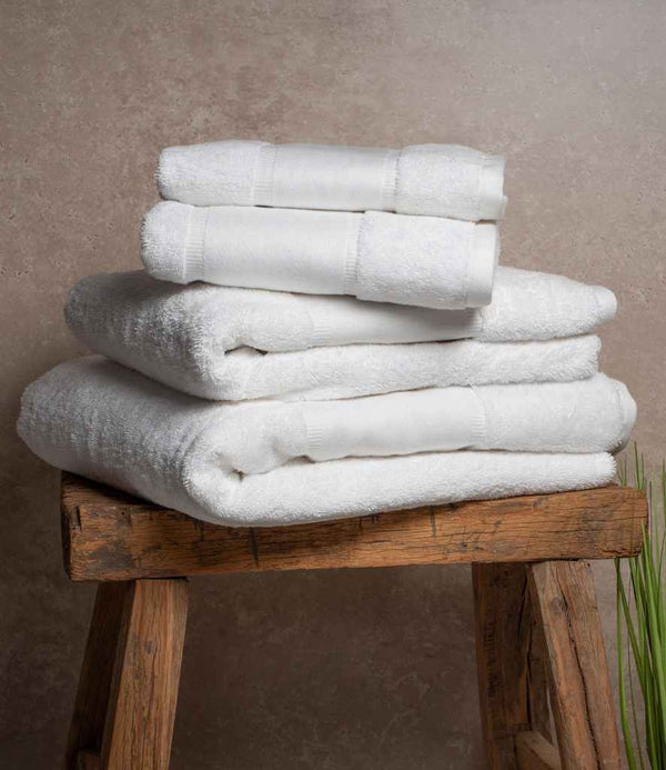 A stack of Towel City Organic Printable Border Guest Towels is neatly arranged on a wooden stool against a neutral textured wall, featuring three large and two small towels, with green plant leaves visible in the lower right corner.
