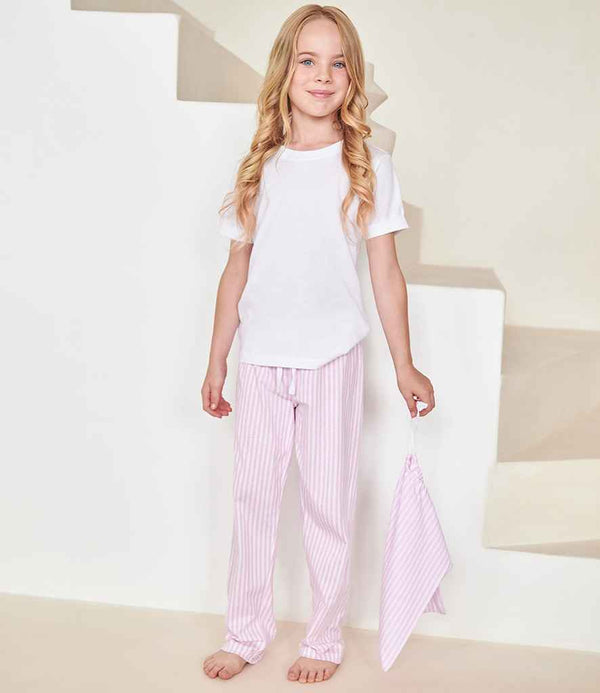 A young girl stands barefoot indoors, wearing Towel City Kids Long PJs by Towel City—a white crew neck top and light pink striped bottoms. She holds a matching striped cloth with stairs in the background.