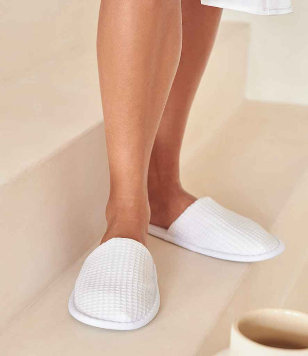 A person wearing a white robe and Towel City Waffle Mule Slippers by Towel City with a mixed fibre sole stands on light stairs. A cup of beverage rests on a lower step in the foreground.