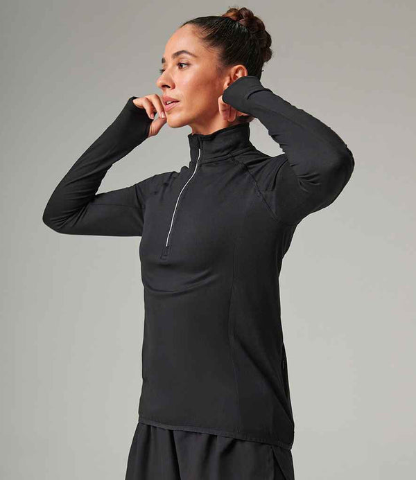 A woman stands sideways against a plain background wearing the Tombo Ladies Long Sleeve Zip Neck Performance Top, adjusting its half-zip collar with both hands. The black athletic top by Tombo features reflective trim.