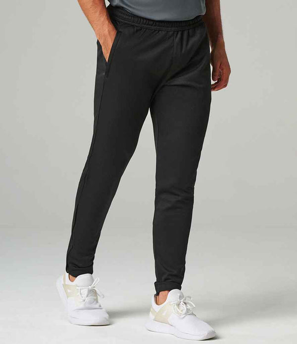 A person wearing Tombo Slim Leg Training Pants by Tombo, paired with a gray shirt and white athletic shoes, stands against a plain light background. Only the lower body is visible.