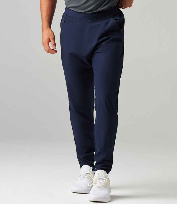 A person models Tombo Technical Trousers—navy blue, tapered leg, elasticated waistband—from Tombo, paired with white sneakers against a plain light gray background. The person’s head is not visible in the image.