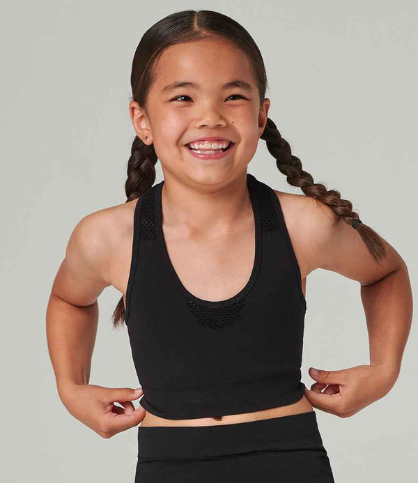 A young girl with braided pigtails smiles while wearing the Tombo Kids Seamless Crop Top by Tombo and matching bottoms, lifting the hem of her top with both hands. The background is a plain light gray.