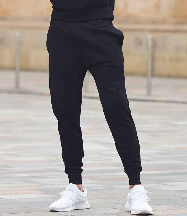 A person stands outdoors in Tombo Unisex Athleisure Joggers by Tombo and white athletic shoes. The image is cropped at the waist, showing only the joggers against a paved area with bollards in the background.