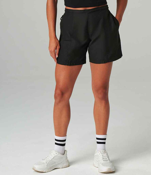 A person wears Tombo Ladies All Purpose Shorts by Tombo, featuring an elasticated waistband and side pockets, paired with a black top and white striped socks and shoes, standing against a plain background.