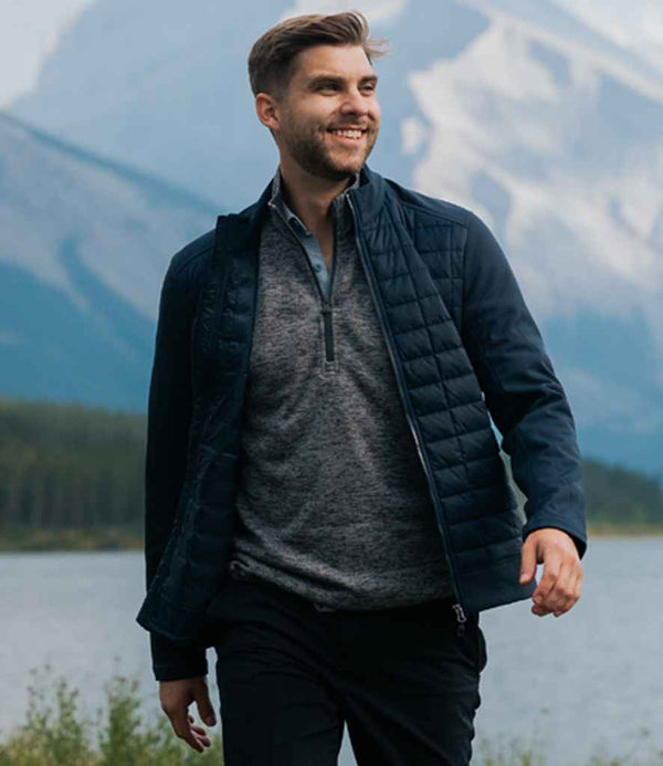 A man with a beard and short hair smiles while walking outdoors by a lake with mountains. He is wearing a Stormtech Appalachian Thermal Soft Shell Jacket over a gray shirt and black pants.