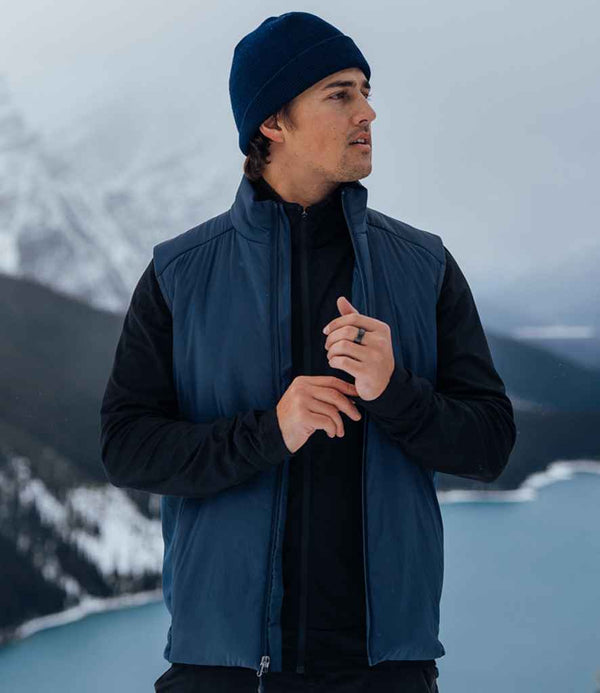 A man wearing a blue Stormtech Cascadia Thermal Bodywarmer and a black long-sleeve shirt stands outdoors by a snowy mountain lake, looking to the side while zipping his vest.