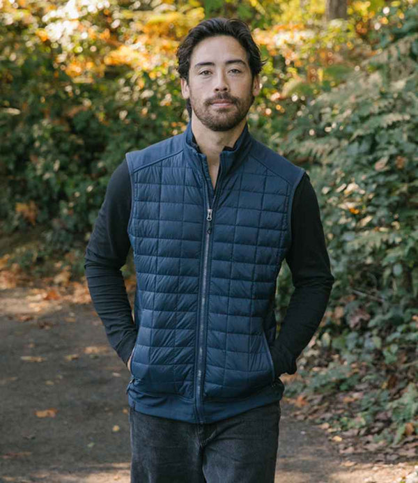 A man with dark hair and a beard stands on a paved path wearing the Stormtech Appalachian Thermal Soft Shell Bodywarmer over a black long-sleeve shirt and dark pants. Green foliage and autumn trees are visible in the background.