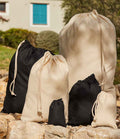 Six Westford Mill Recycled Cotton Stuff Bags in black and beige, with secure drawcord closures, are displayed outdoors on rocks with greenery and a building featuring a blue window in the background.