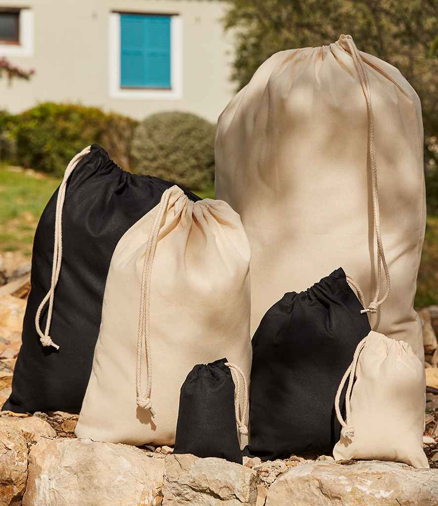 Six Westford Mill Recycled Cotton Stuff Bags in black and beige, with secure drawcord closures, are displayed outdoors on rocks with greenery and a building featuring a blue window in the background.