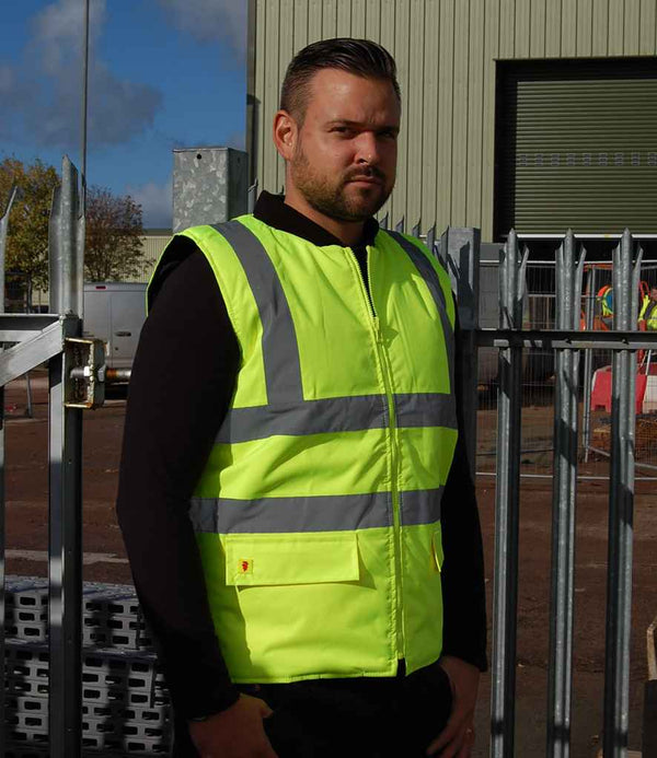 A man wearing a Warrior Hi-Vis Reversible Bodywarmer by Warrior stands in front of a metal fence at an industrial site, with a warehouse and construction materials visible in the background.