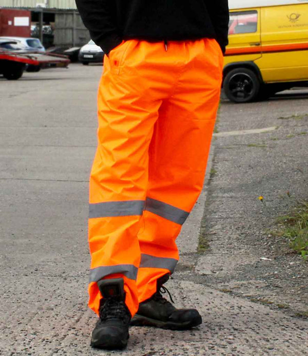A person stands outside wearing Warrior Hi-Vis Overtrousers—bright orange, waterproof, EN ISO 20471 compliant with reflective bands—from the brand Warrior, along with black work boots. In the background are a yellow vehicle and a black-and-red car.
