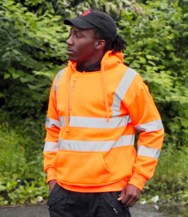 A person wearing the Warrior Hi-Vis Pull On Hoodie by Warrior, which meets EN ISO 20471 standards and features reflective stripes, stands outdoors in front of green foliage, looking to the side with hands in their pockets and a black cap.