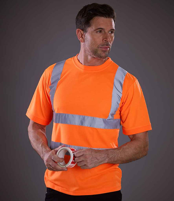 A man wearing a YOKO Hi-Vis Short Sleeve T-Shirt with EN ISO 20471 reflective bands stands against a plain background, holding a roll of tape in both hands.