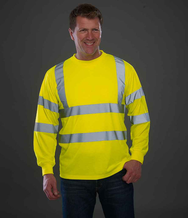 A man stands against a gray background, smiling with one hand in his pocket, wearing the YOKO Yoko Hi-Vis Long Sleeve T-Shirt featuring bright yellow fabric and reflective bands. This EN ISO 20471 class 3 shirt ensures high visibility.