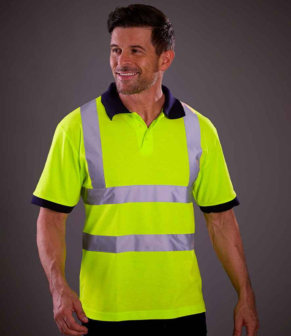 A man wears the YOKO Hi-Vis Polo Shirt with reflective silver stripes, certified to EN ISO 20471 class 2. He stands against a dark background, looking to the side and smiling.