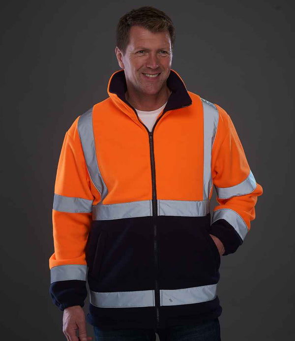 A man smiles with one hand in his pocket, wearing the YOKO Yoko Hi-Vis Heavyweight Fleece Jacket in orange/navy with EN ISO 20471 class 3 reflective bands, standing against a dark gray background.