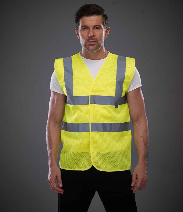 A man faces the camera wearing a Yoko Top Cool Open Mesh Waistcoat by YOKO, featuring a bright yellow mesh design and reflective bands that meet ANSI/ISEA 107 standards, over a white T-shirt, against a plain gray background.