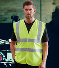 A man with short brown hair and a beard wears a YOKO Yoko Hi-Vis Anti-Static Flame Retardant Waistcoat with silver reflective stripes, meeting EN ISO 20471 standards, over a black shirt. He stands indoors against a blurred industrial background.