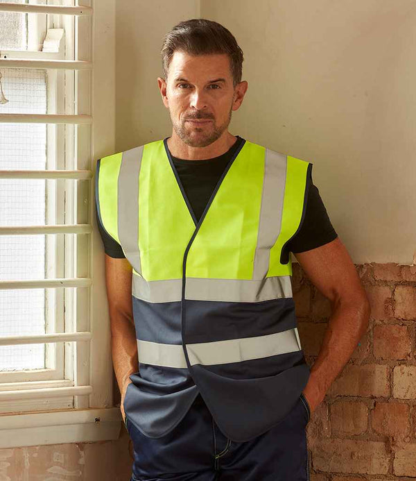 A man wearing a YOKO Two Tone Waistcoat, a high visibility reflective vest compliant with EN ISO 20471 standards, stands indoors leaning against a windowsill beside an exposed brick wall.