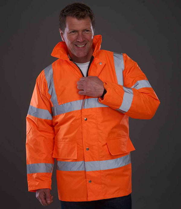 A man smiles as he adjusts a YOKO Yoko Hi-Vis Classic Motorway Jacket in bright orange with silver stripes. The EN ISO 20471 waterproof jacket has a high collar, snap buttons, and two large front pockets, set against a plain gray background.