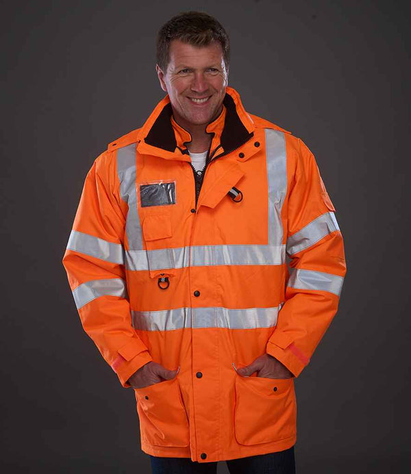 A man stands against a dark background, smiling at the camera with his hands in the pockets of a bright orange YOKO Hi-Vis Multi-Function 7-in-1 Jacket, featuring reflective silver stripes and made by YOKO.