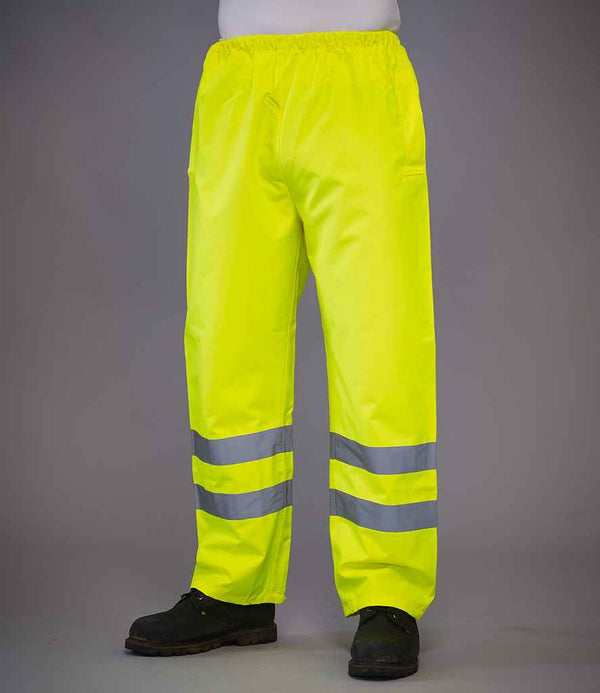 A person wearing YOKO Yoko Hi-Vis Waterproof Overtrousers in bright yellow with two horizontal reflective ankle bands and black shoes stands against a plain gray background.
