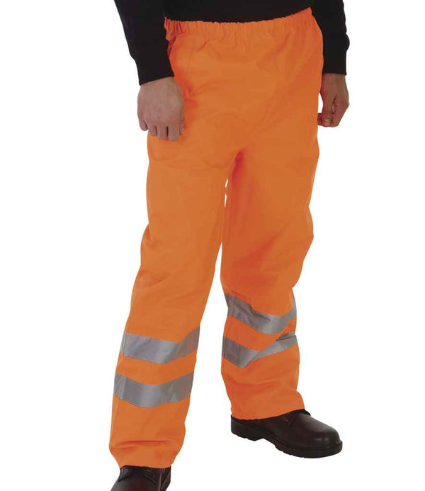 A person wearing YOKO Yoko Hi-Vis Waterproof GO/RT Overtrousers in bright orange with two silver reflective bands near each legs bottom, paired with black shoes and a black long-sleeve shirt.