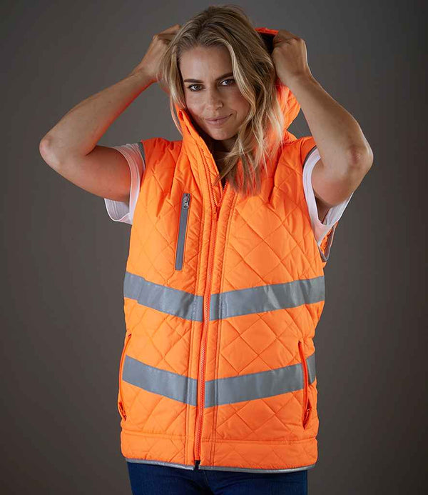 A person wearing the Yoko Hi-Vis Kensington Hooded Gilet by YOKO, featuring bright orange color, reflective bands, and a hood, stands against a plain gray background holding the hood with both hands.