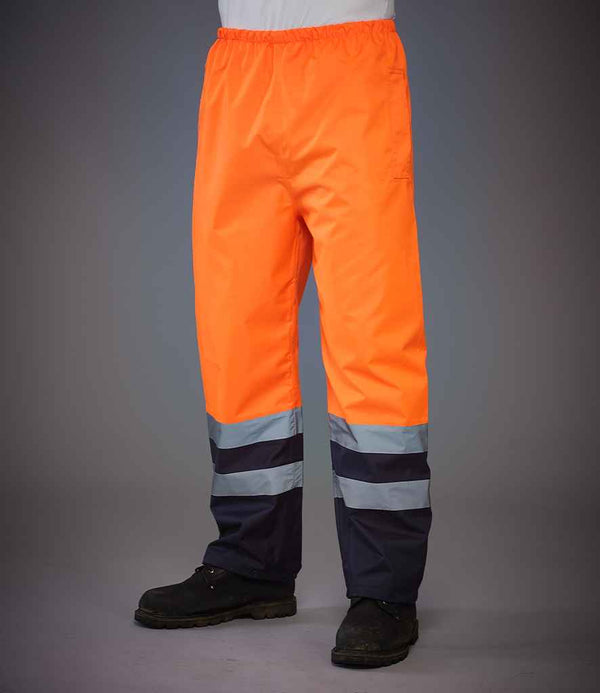 A person wears YOKO Yoko Hi-Vis Two Tone Waterproof Overtrousers in bright orange with reflective stripes near the cuffs and black safety boots, shown from the waist down against a plain gray background.