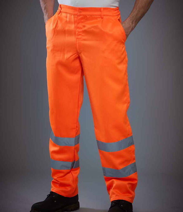 A person is seen from the waist down wearing YOKO Hi-Vis Poly/Cotton Work Trousers in bright orange with water repellent fabric and reflective silver bands, along with black shoes and a white shirt.