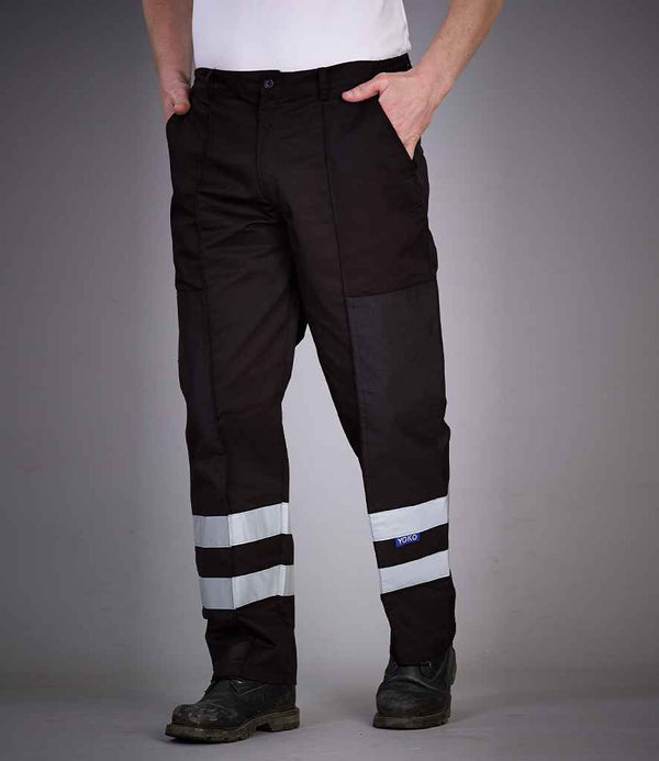A person wearing YOKO Yoko Reflective Poly/Cotton Ballistic Trousers with two horizontal reflective bands near the bottom of each leg, black shoes, and a white shirt. Their hands are in their pockets and their face is not visible.