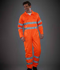 A person wearing the YOKO Hi-Vis Poly/Cotton Coverall stands against a dark background. This bright orange coverall features reflective bands, silver stripes, is EN ISO 20471 compliant, and water repellent for added protection.