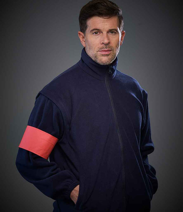 A man with short brown hair and stubble wears a navy zip-up jacket and a YOKO Yoko Print Me Arm Band in high-visibility red on his left arm, standing against a dark gray background with his hands in his pockets.
