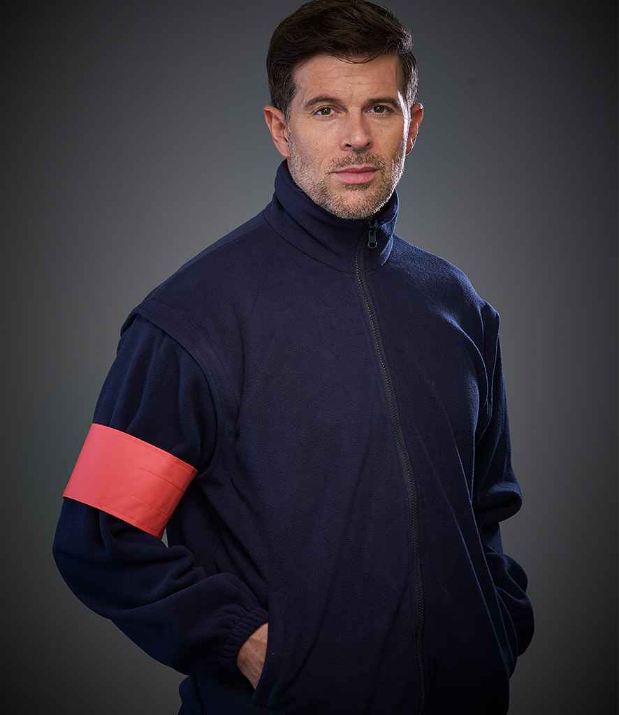 A man with short brown hair and stubble wears a navy zip-up jacket and a YOKO Yoko Print Me Arm Band in high-visibility red on his left arm, standing against a dark gray background with his hands in his pockets.